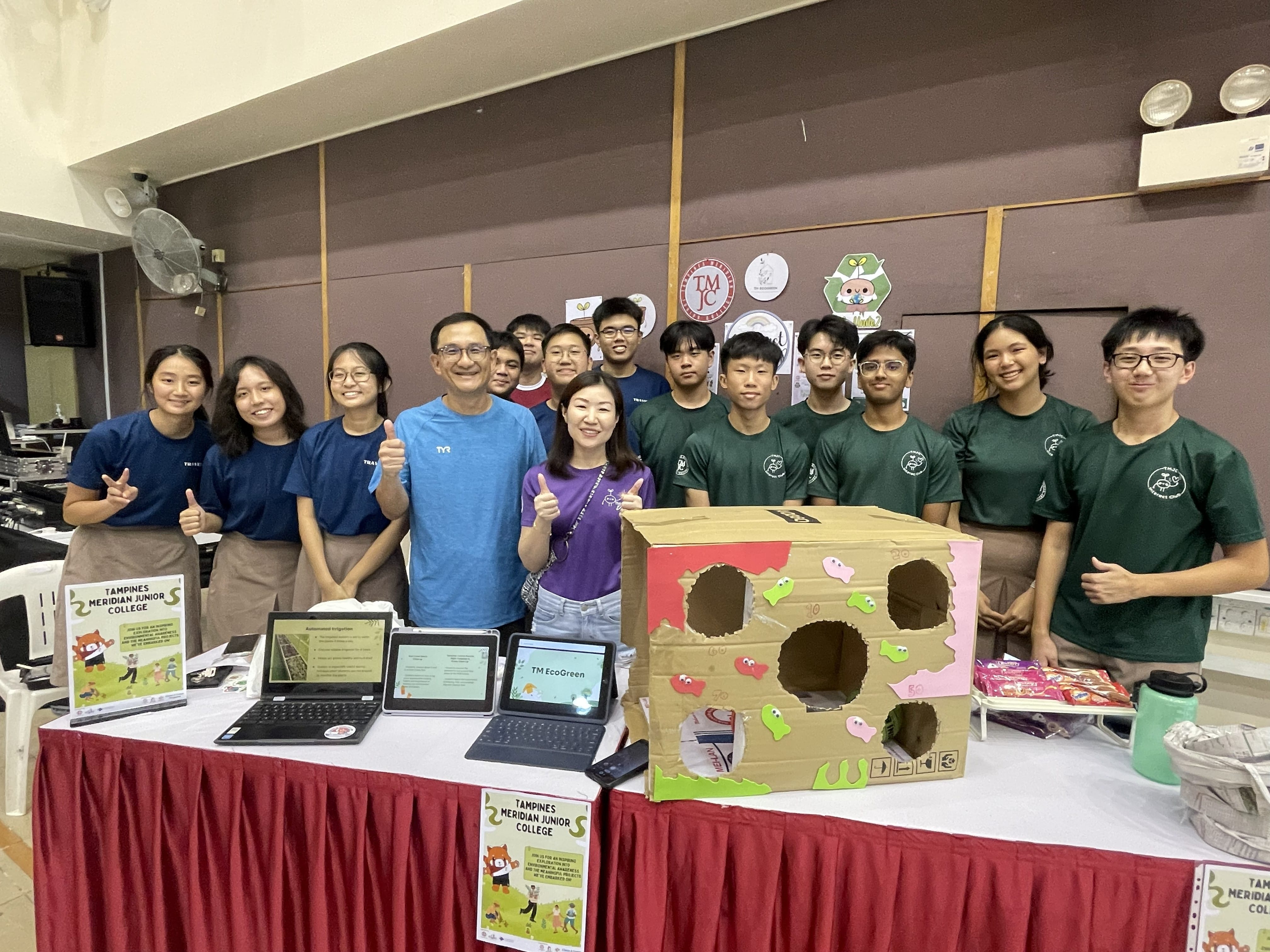 A large group of TMJC Interact Club students and two adult guests pose smiling behind a booth displaying tablets, posters, and a large decorated cardboard box as part of their "TM EcoGreen" environmental outreach at the Sengkang North Eco Fest.